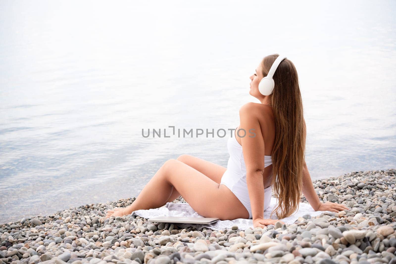 A woman is sitting on a beach with her headphones on. She is wearing a white bikini and a white shirt. The beach is rocky and the water is calm. The woman is enjoying her time by the water