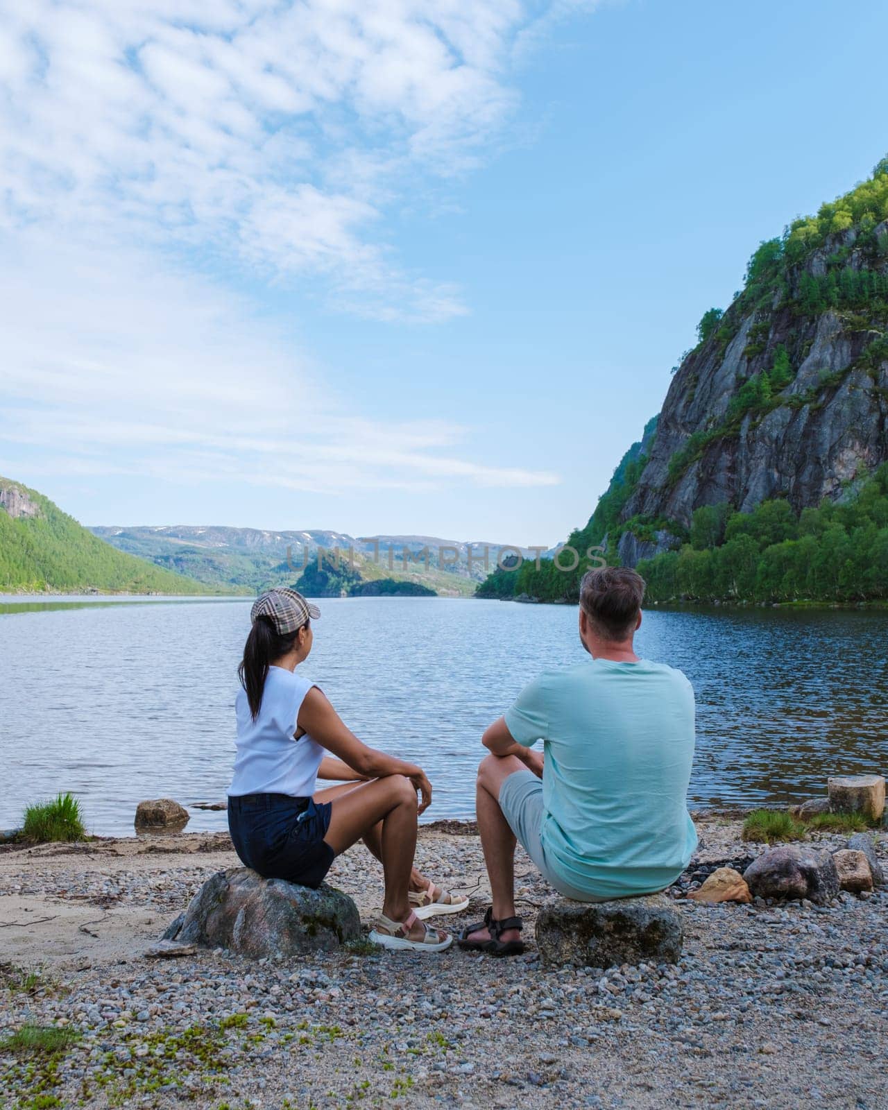 A couple sits on rocks by a tranquil lake in Norway, enjoying the breathtaking scenery and quiet solitude.