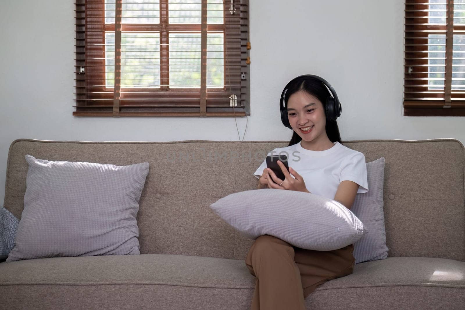 A woman sitting on a sofa, wearing headphones and using her smartphone, symbolizing modern relaxation and digital entertainment in a cozy home setting