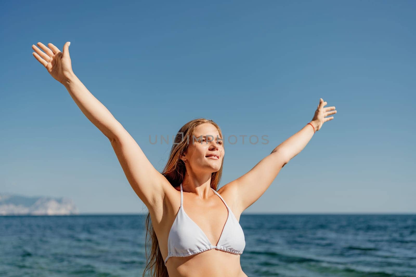 A woman in a bikini is standing on the beach and is smiling. She is reaching out with her arms open, as if she is embracing the ocean. Concept of joy and relaxation