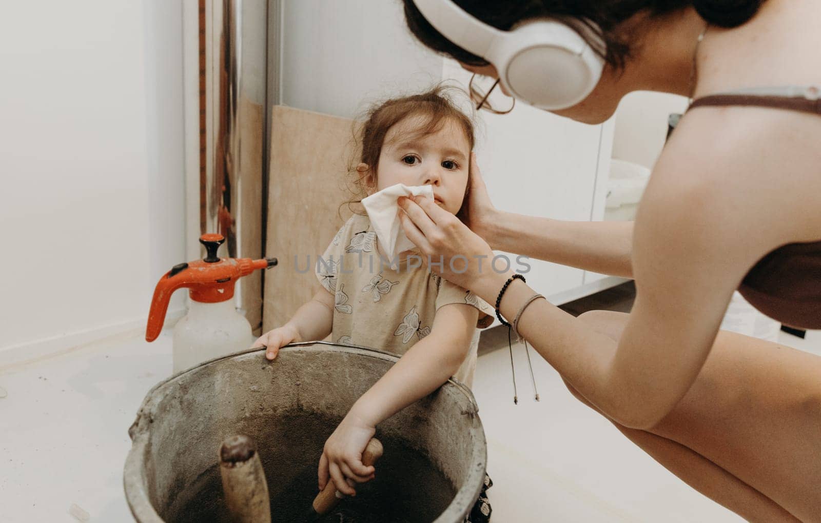 One Caucasian teenage girl wipes the dirty mouth of her younger beautiful sister with a napkin, who mixes cement mortar in a bucket with a spatula, in a room where renovation and installation of a smoker for a fireplace made of bricks is underway, bottom side view close-up with selective focus. Concept of clean brickwork.