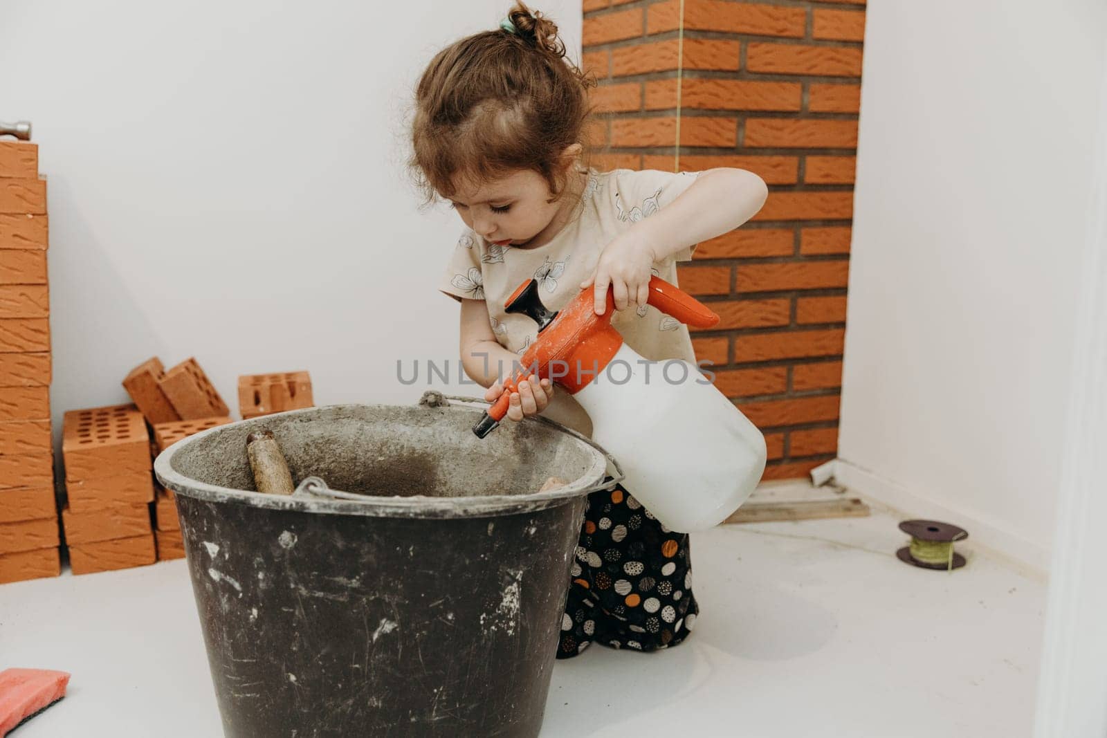 One small beautiful Caucasian girl sits on her knees and uses a hand sprayer to wet cement mortar in a bucket, in a room where repairs and installation of a smoker for a fireplace made of bricks are underway, close-up side view with selective focus. Concept of clean brickwork.