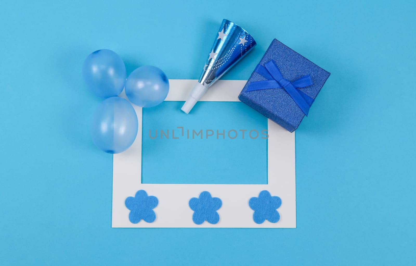 Empty paper frame, three inflated mini balloons, one whistle, decorative felt flowers and a shiny gift box lie on a blue background with copy space in the center, flat lay close-up.