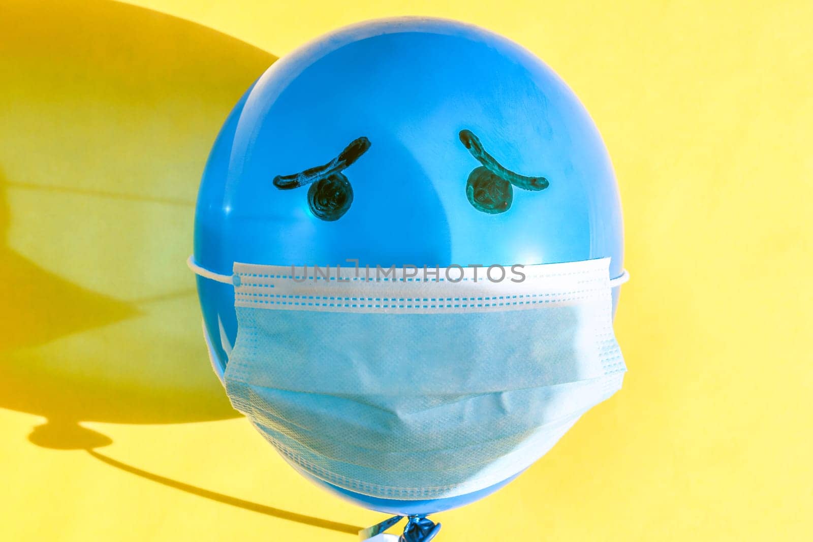 One blue balloon with a medical mask and painted black sad eyes lies in the center on a yellow background on a summer sunny day with hard shadows on a yellow background, flat lay close-up.