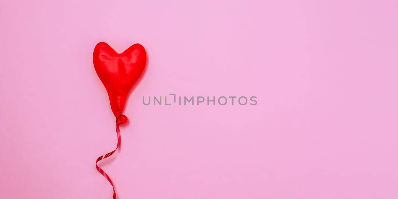 One red deflated heart-shaped balloon lies in the left on a pink background with copy space on the right, flat lay close-up.