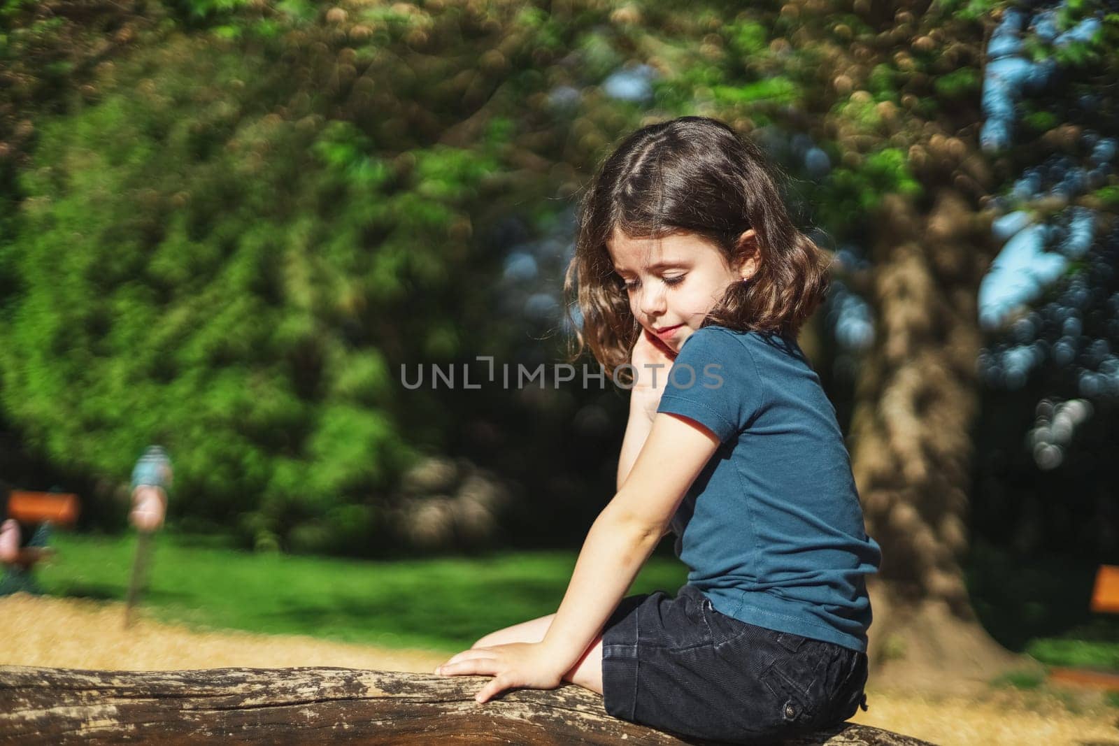 Portrait of a caucasian beautiful girl with short brown flowing hair, a blue t-shirt and shorts sits half-side on a wooden pole from a rope swing in a park on a playground, close-up side view with selective focus. The concept of PARKS and REC, happy childhood, children's picnic, holidays, children's recreation, outdoors.