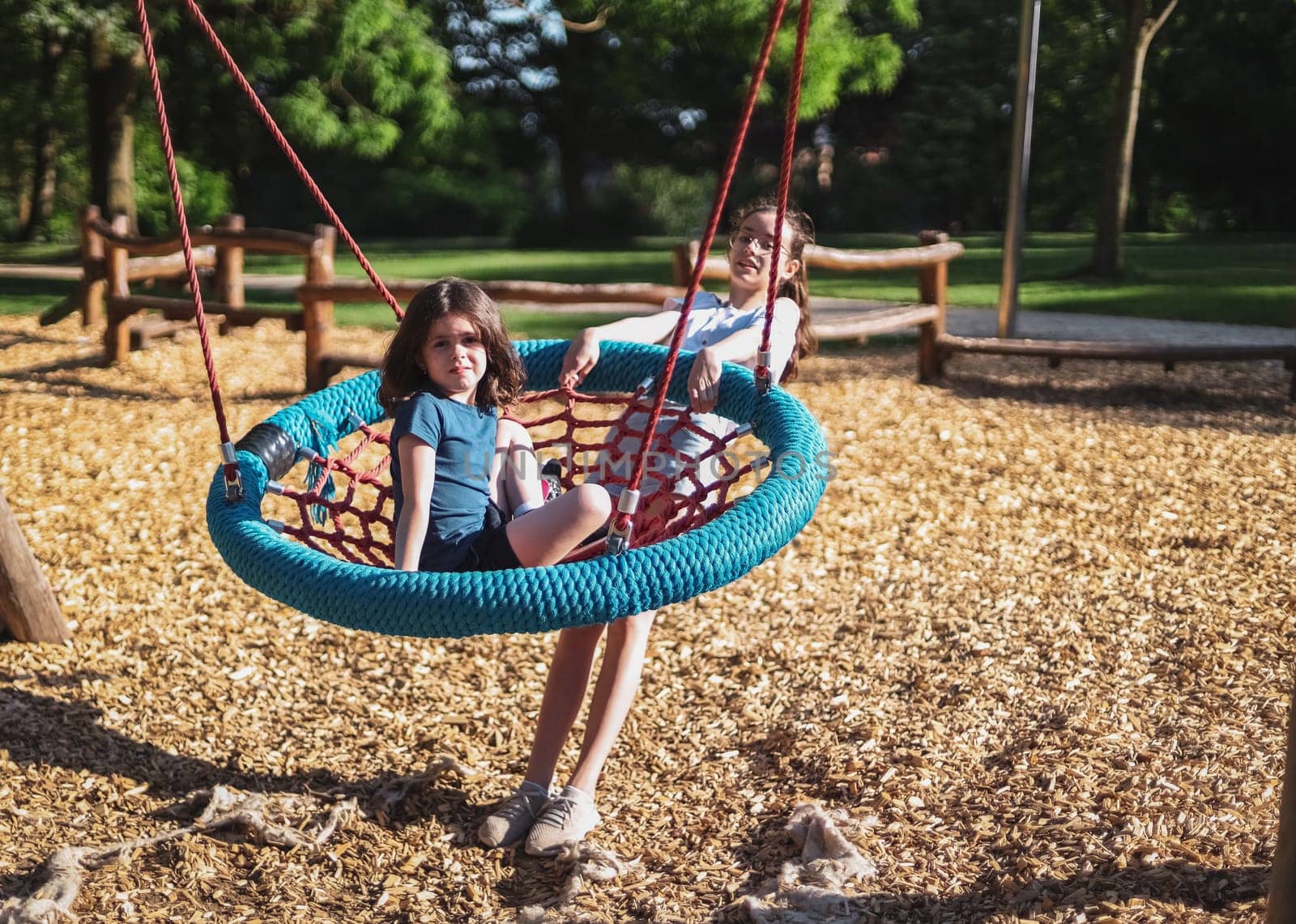 Portrait of two beautiful caucasian girls sisters: the older one swings the younger one on a round rope swing in the park on the playground, close-up side view. The concept of PARKS and REC, happy childhood, children's picnic, holidays, children's recreation, outdoor recreation, playgrounds, outdoors, family recreation.