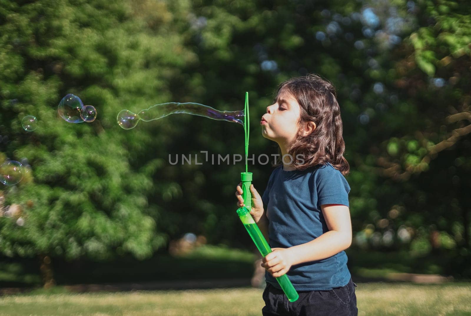 Portrait of one beautiful Caucasian brunette girl blowing a long soap bubble, standing on the right in the park on the playground, close-up side view. The concept of PARKS and RECREATION, happy childhood, children's picnic, happy childhood, outdoor recreation, playgrounds.