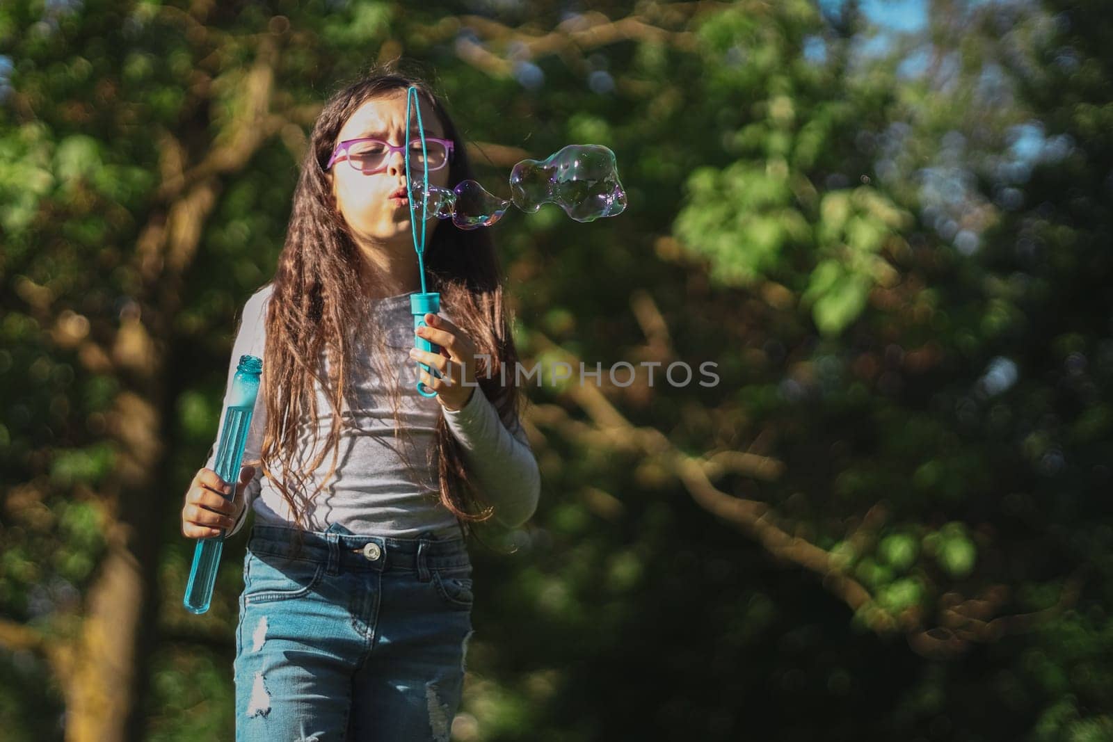 Portrait of one beautiful Caucasian brunette girl in glasses emotionally blowing bizarre shapes of soap bubbles, standing on the left in the park on the playground, close-up view from below. Concept of PARKS and RECREATION, happy childhood, children's picnic, happy childhood, outdoor recreation, playgrounds.