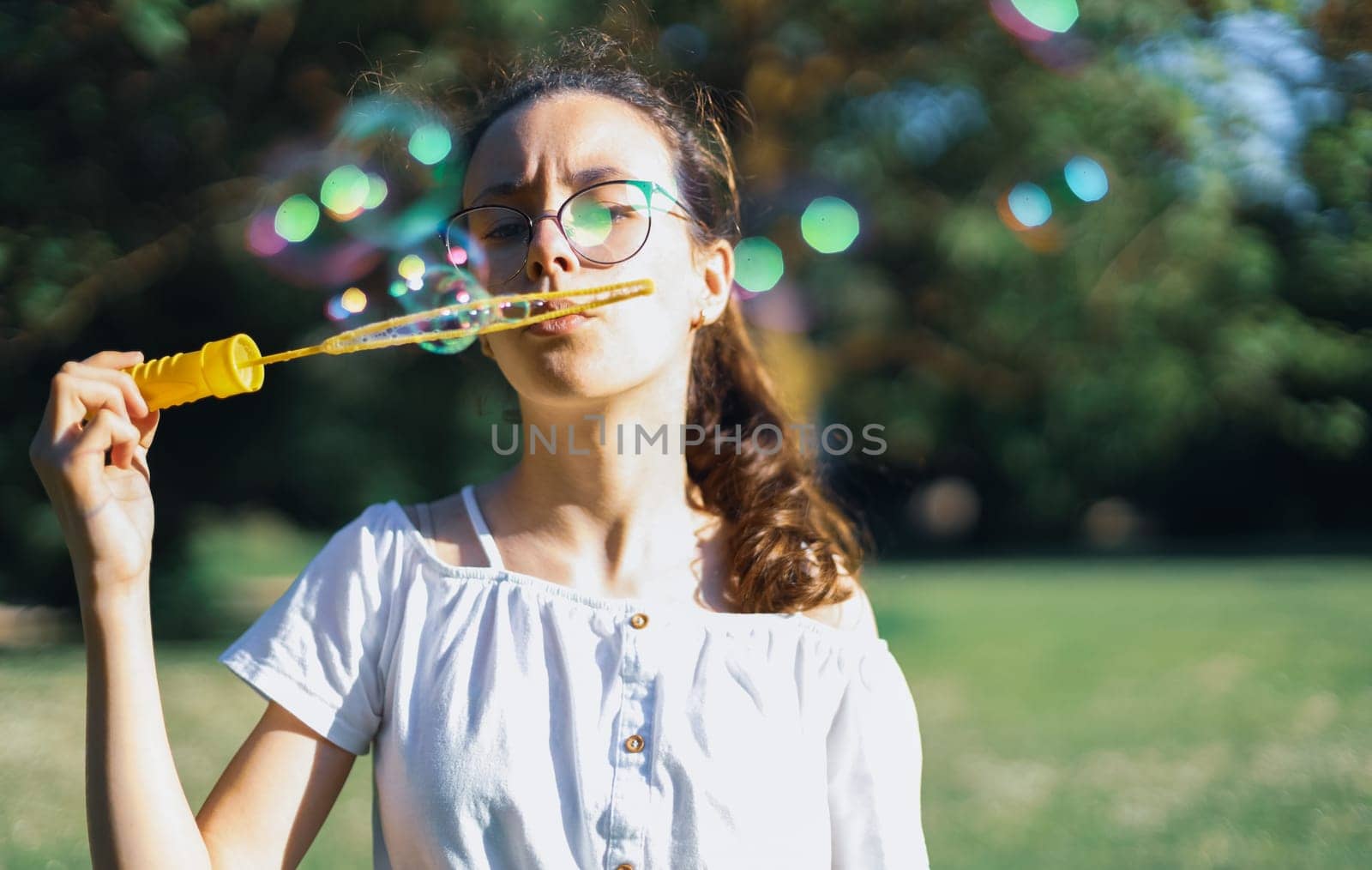 Portrait of one beautiful Caucasian teenage girl in glasses with closed eyes blowing soap bubbles, standing in the park on the playground, close-up side view with selective focus. Concept of PARKS and RECREATION, happy childhood, children's picnic, happy childhood, outdoor recreation, playgrounds.