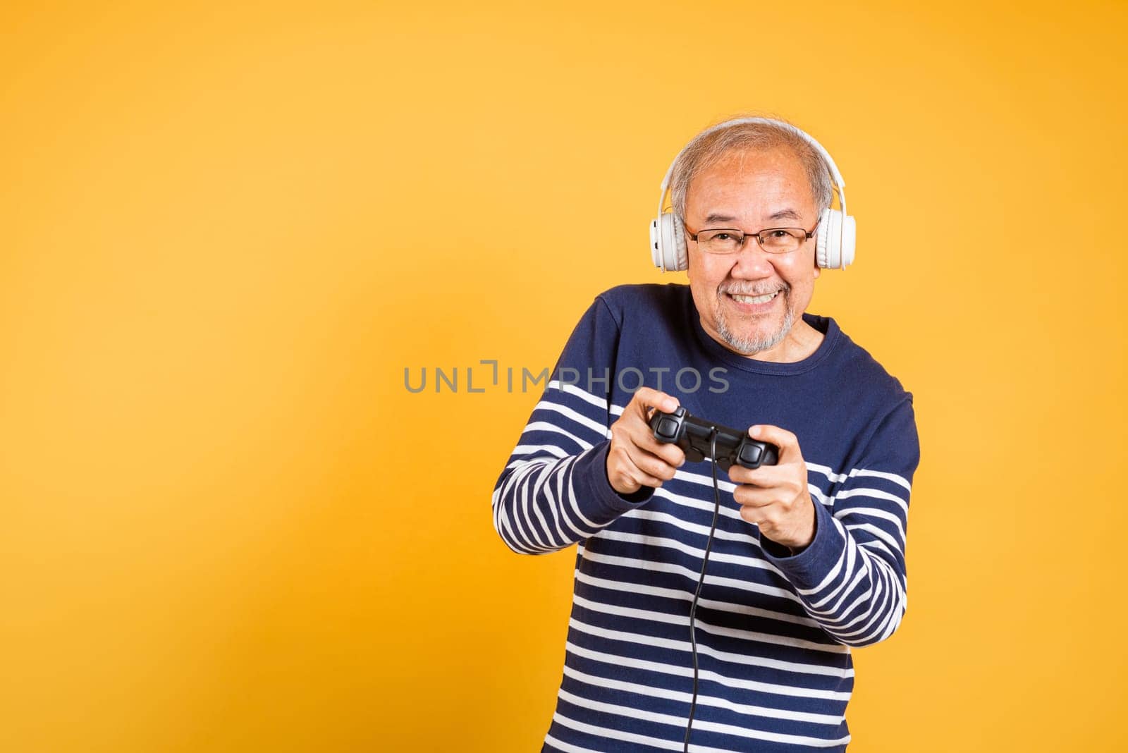 Portrait Asian smiling old man with headphones holding game controller studio shot isolated yellow background, senior man pensioner feeling winning playing a video game with joystick console