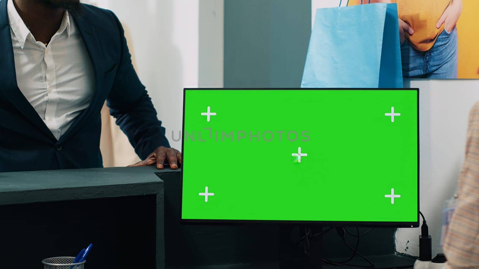 African american worker uses computer with greenscreen display on at checkout counter, putting clothes in a bag for customer. Employee looks at pc showing copyspace mockup screen. Camera A.