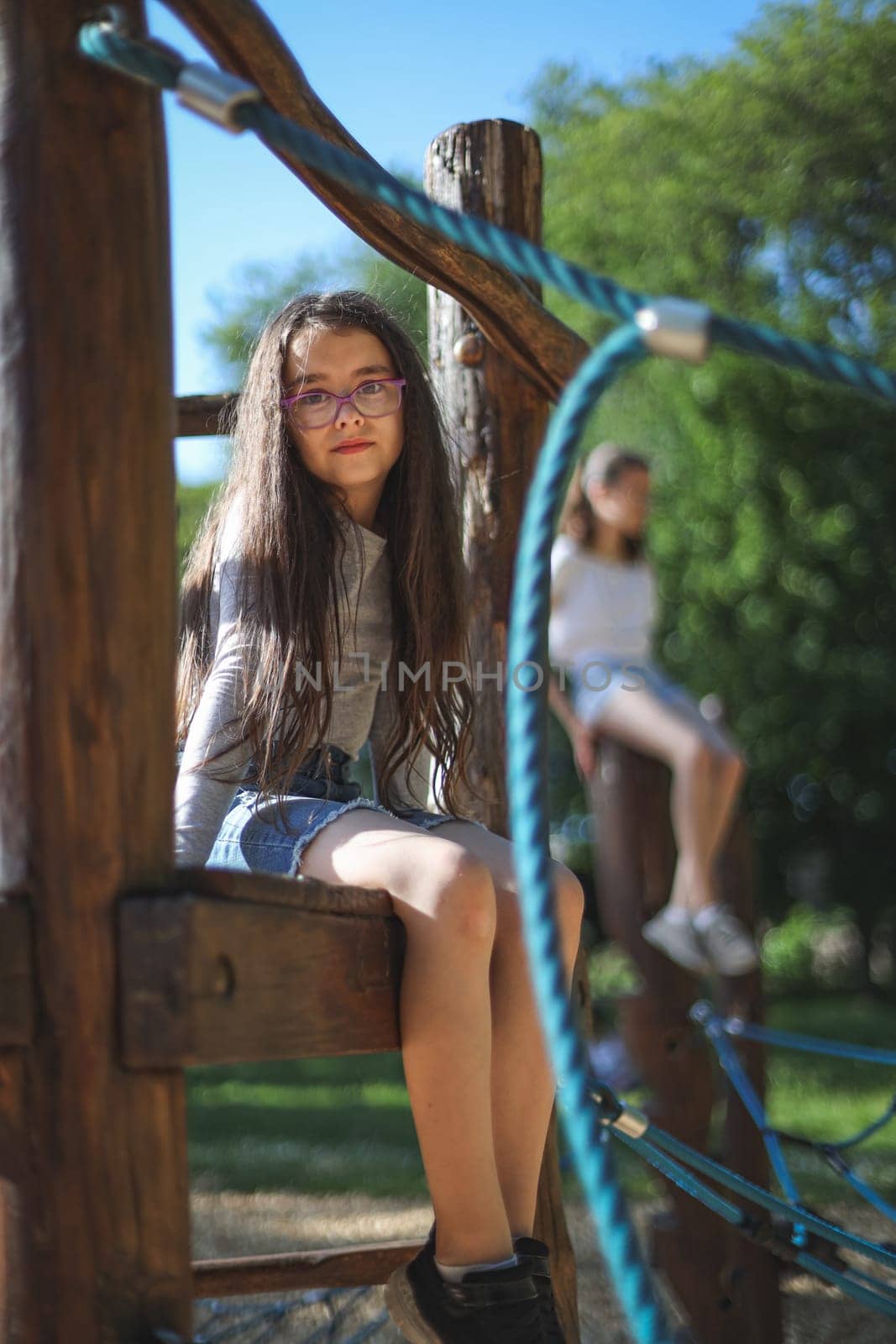 Portrait caucasian girl with long flowing brown hair, glasses in a gray long sleeve t-shirt sits on a rope swing looking at the camera in a park on a playground, close-up bottom view with selective focus and blurred background. The concept of PARKS REC, happy childhood, children's picnic, holidays, children's recreation, outdoor recreation, children entertainment, outdoor activities.
