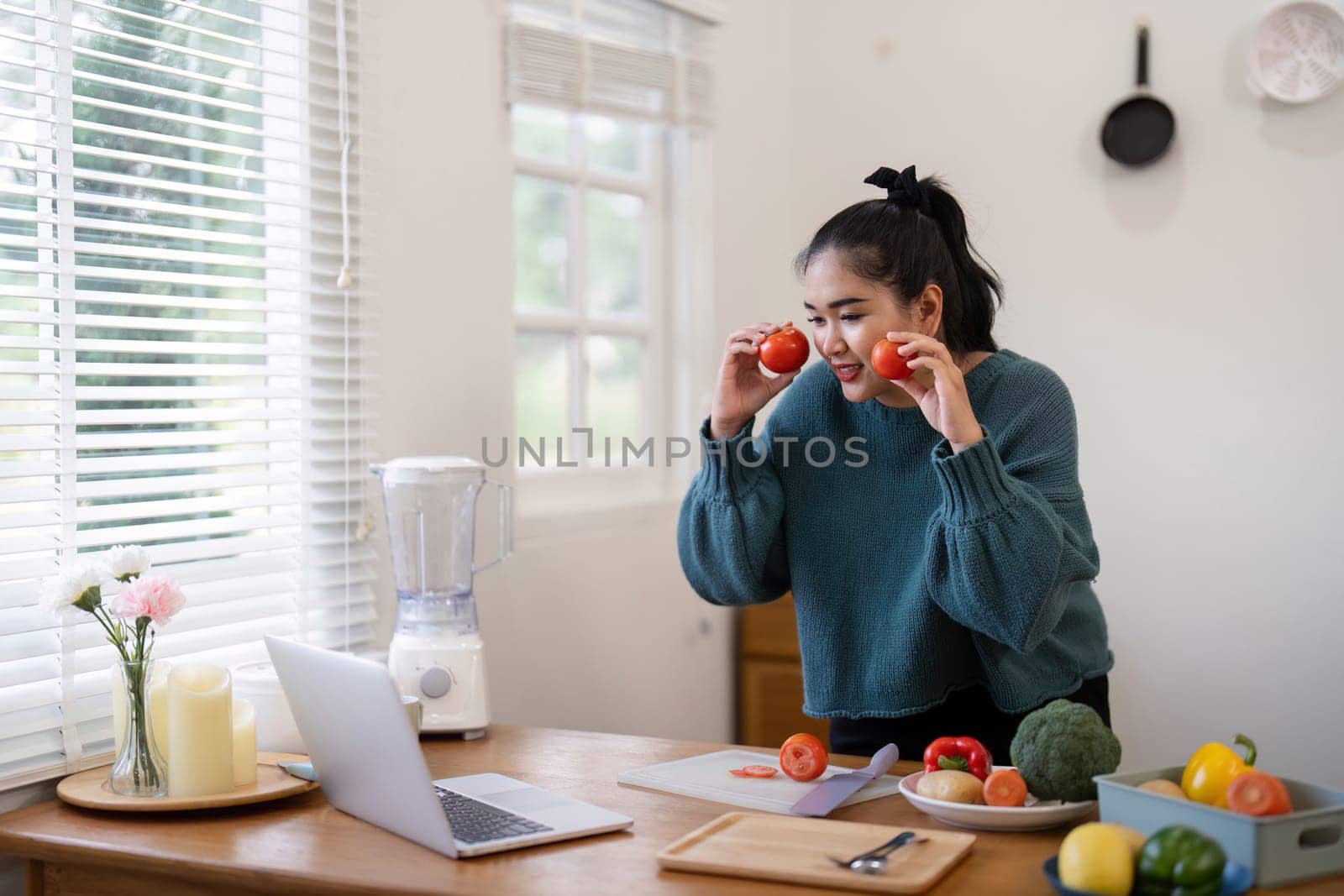 Young woman in a kitchen holding tomatoes, preparing vegetables on a cutting board, and video chatting via laptop, highlighting healthy cooking and online communication