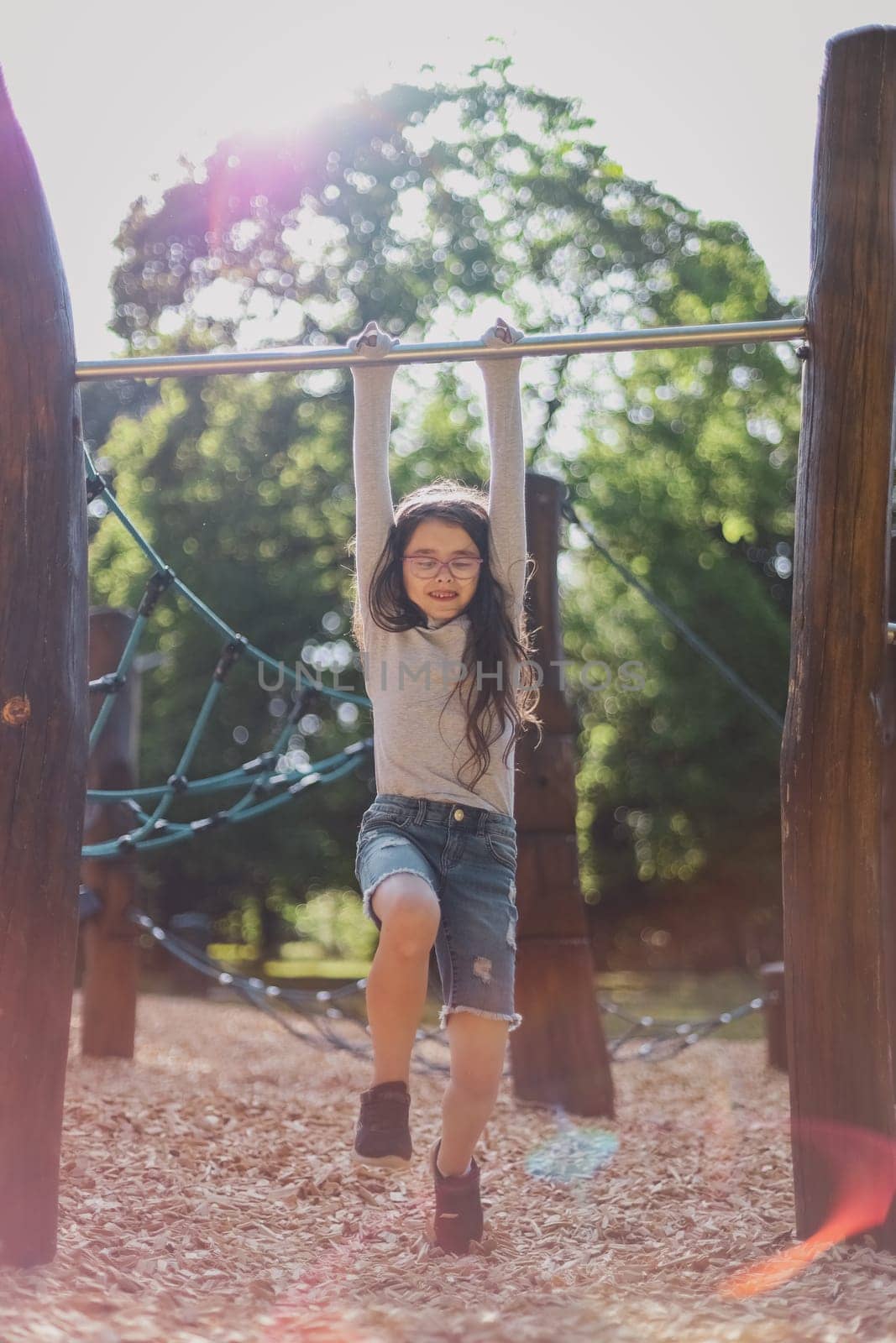 Portrait of a beautiful caucasian girl with long flowing brown hair in a gray long-sleeve T-shirt, shorts and glasses is doing push-ups on the horizontal bar in the park on the playground in sun glare, close-up view from below with selective focus. The concept of PARKS and REC, happy childhood, children's picnic, holidays, fabulous childhood, outdoor recreation, playgrounds, outdoors, push-ups on the horizontal bar, sports games, children camp.