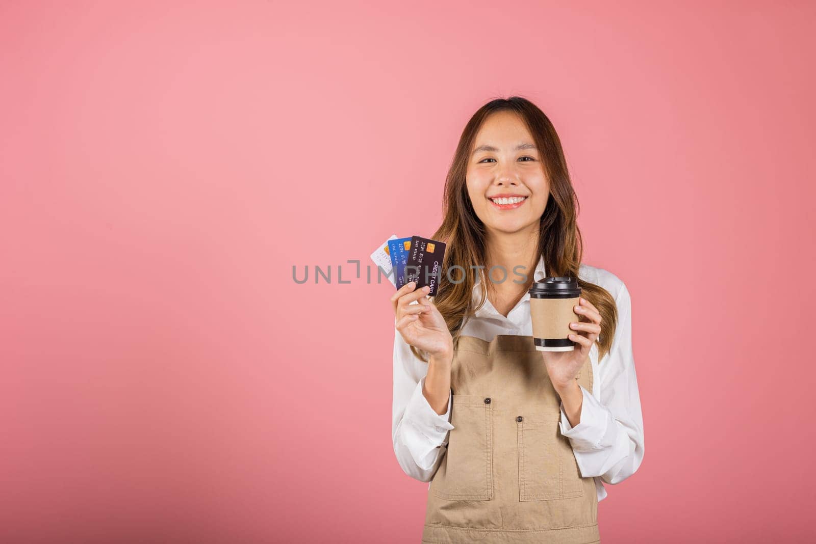 Portrait Asian happy young woman barista bar tender coffee maker holding coffee tea hot cup and credit card studio shot isolated on pink background, female smiling hold takeaways cup for payment