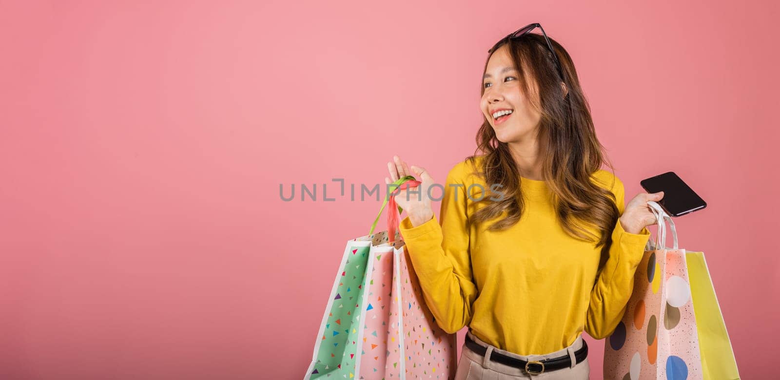 Portrait Asian happy beautiful young woman teen shopper smiling standing excited holding online shopping bags online colorful on mobile phone or smartphone studio shot isolated on pink background