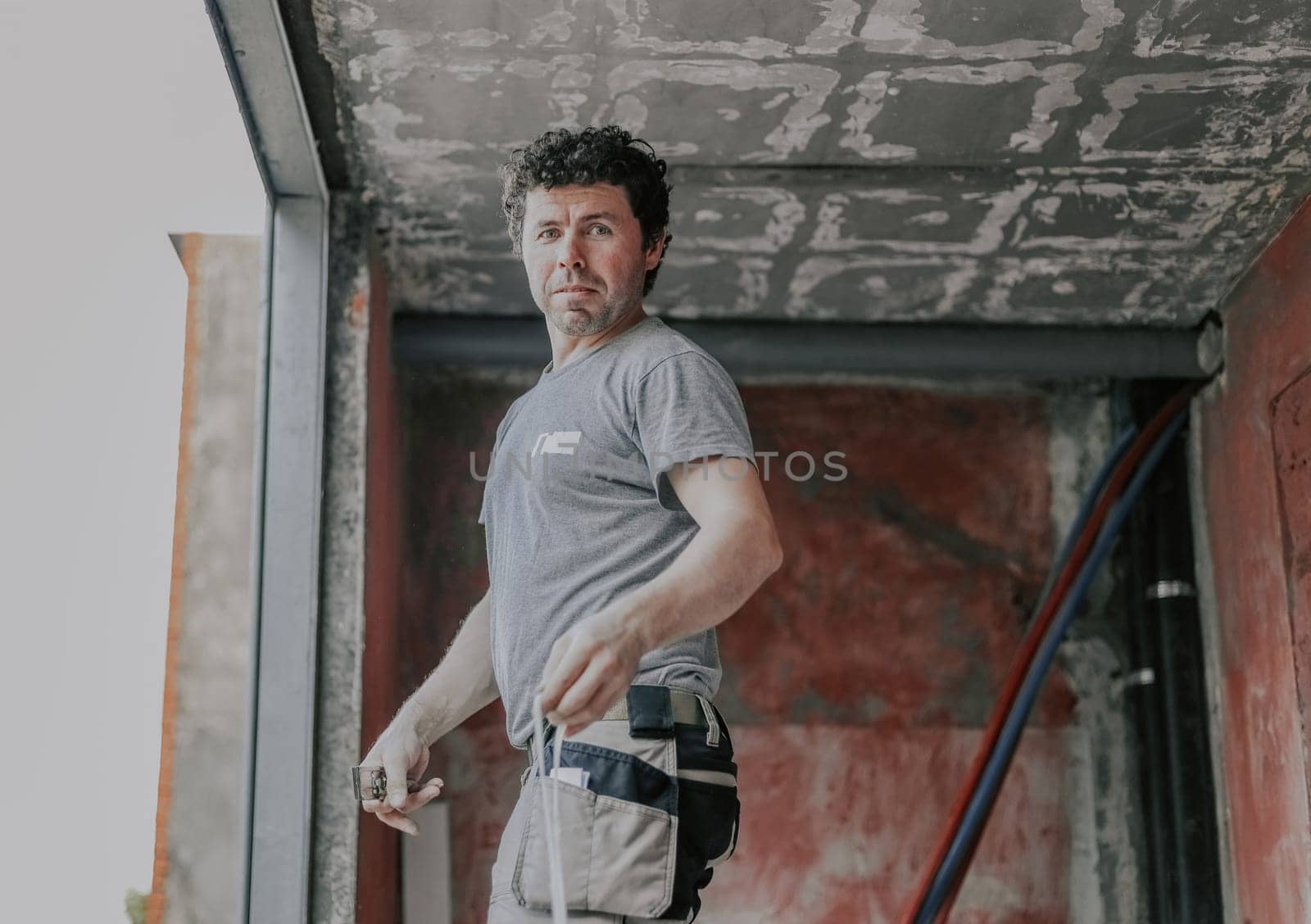 A young caucasian man in a gray t-shirt with curly brown hair stands on a stepladder and looks smiling at the camera, prepares to set the window, close-up view from below with depth of field. The concept of installing windows, construction work, house renovation.