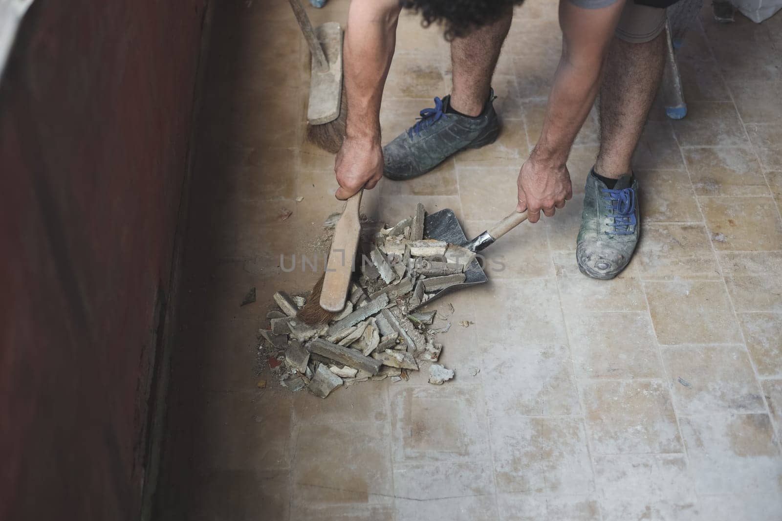 Young caucasian unrecognizable man sweeping construction debris with a small whisk on an old dustpan from a dirty tiled floor, close-up side view. The concept of cleaning and installing windows, construction work, house renovation.