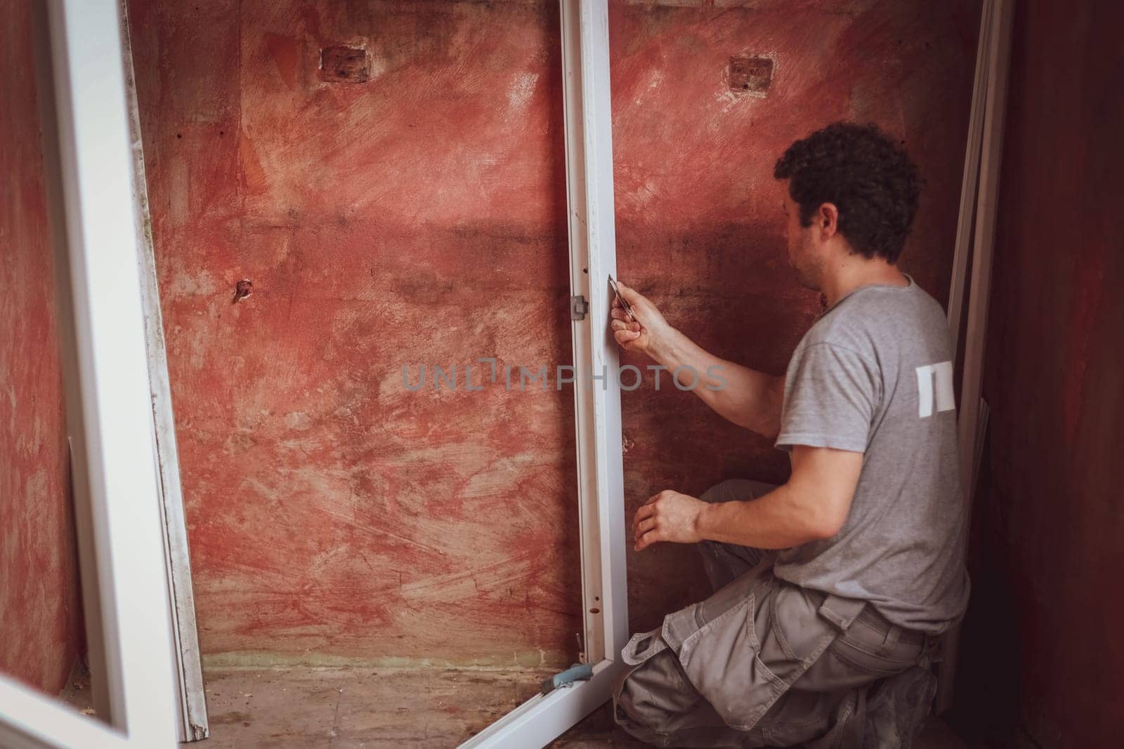 A handsome young Caucasian man with curly brown hair in a gray t-shirt and shorts washes white window frames with a sponge and soap against a red wall, preparing them for installation, close-up side view. The concept of home renovation, window installation, construction work, diy, lifestyle, at home, plastic cleaning, cleaning services, small business, private services.