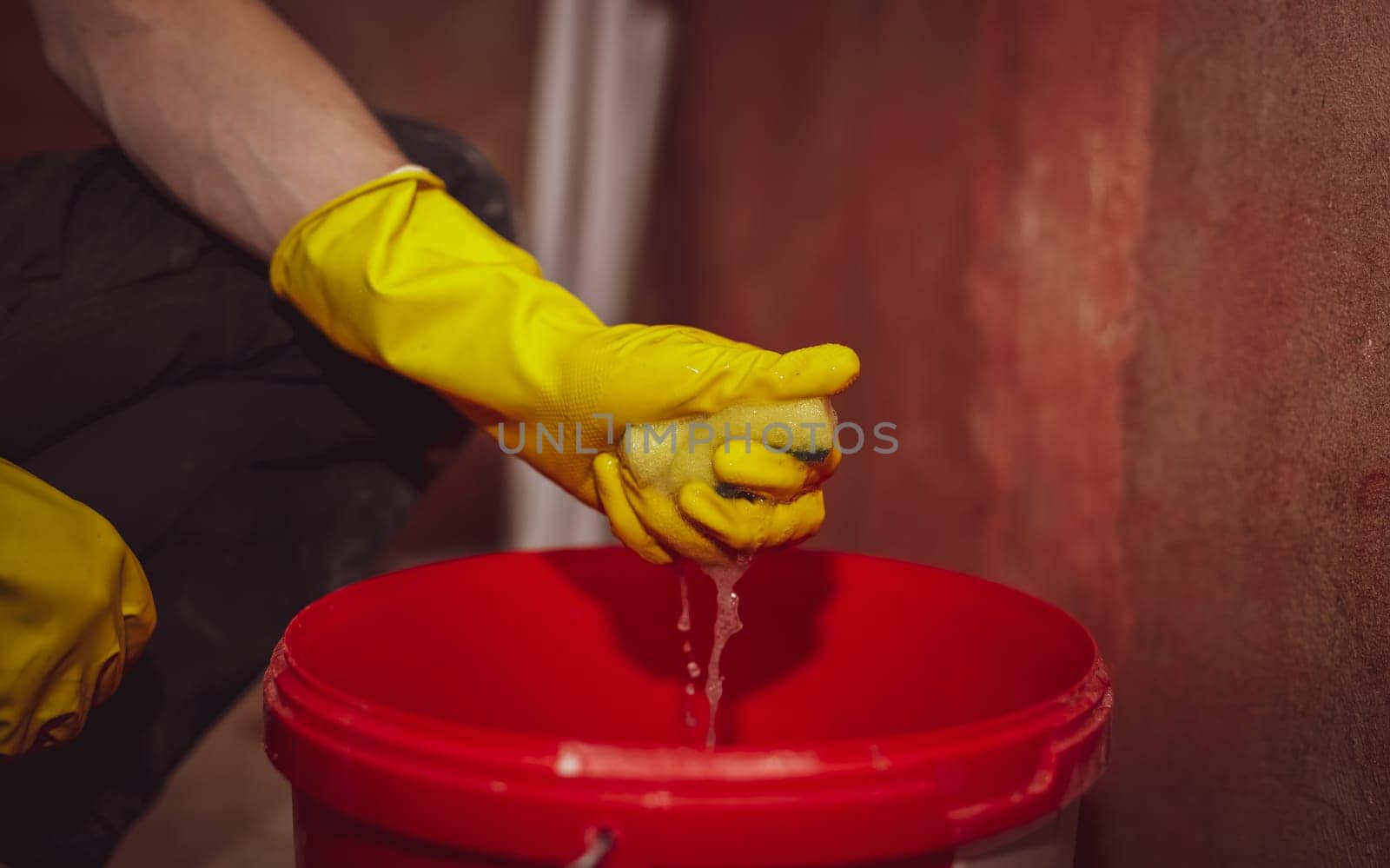 Young caucasian man in yellow gloves wringing out a sponge from the water over a red bucket, close-up side view. The concept of home renovation, window cleaning, construction work.