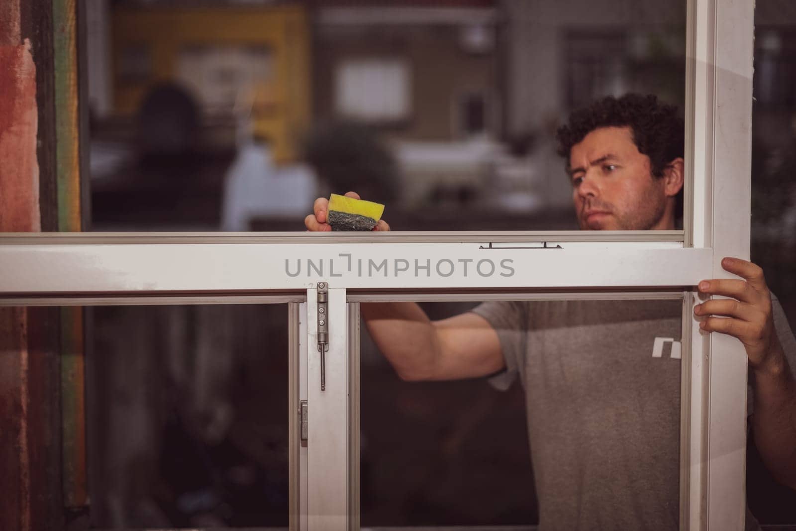 Handsome young caucasian satisfied man washing a window frame without glasses with a sponge and soap, preparing them for installation against a red wall, side view close-up. The concept of home renovation, installation of windows, construction work.