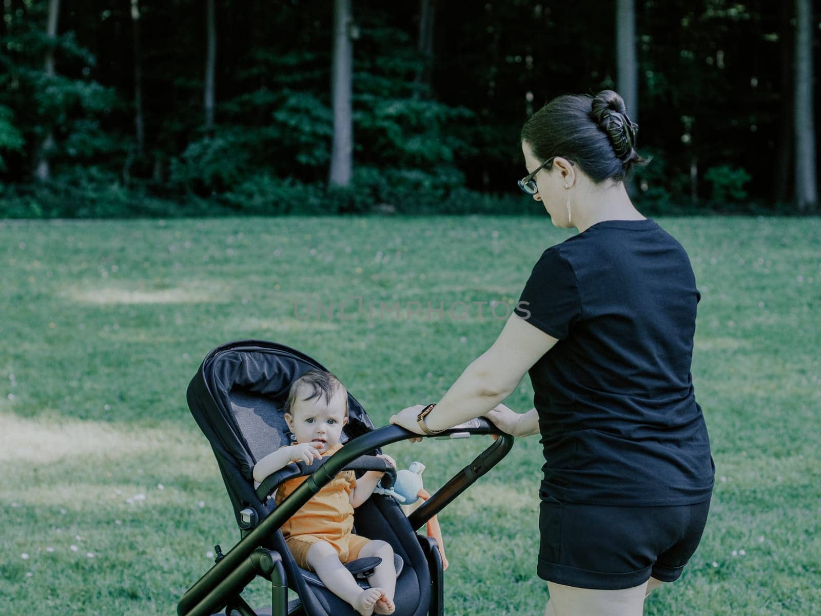 Caucasian young and beautiful woman mother in a black t-shirt and shorts cockles in her little daughter's stroller on a green meadow in a public park against the backdrop of blurry trees, side view close-up. The concept of motherhood, moms, family vacation, spring walk, mother care.