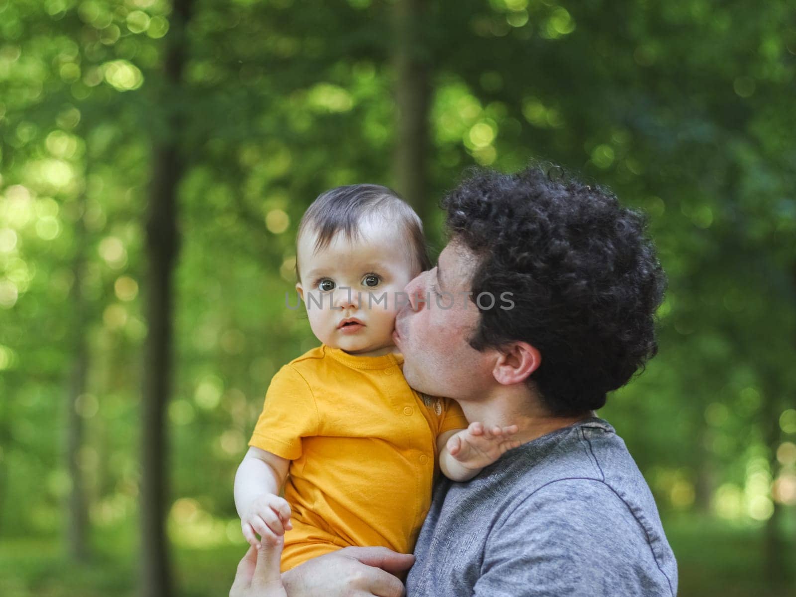 Caucasian young and handsome dad in a blue t-shirt with brown curly hair holds his little daughter in yellow overalls and gently kisses her with love on the cheek, while she looks into the camera in a public park against the backdrop of green trees, close-up side view. The concept of fatherhood, dads, family vacation, spring walk, happy childhood, park and rec.