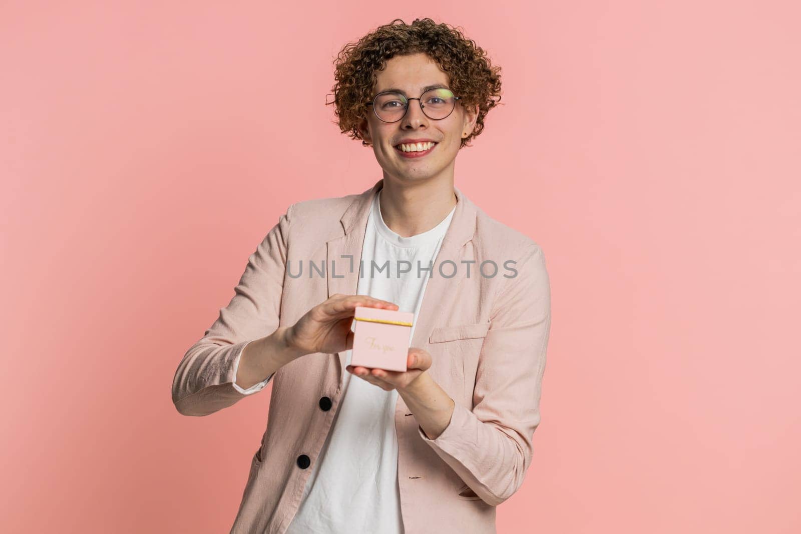 Smiling happy Caucasian man presenting jewelry gift box stretches out hands offer wrapped present career bonus celebrating party promotion discount sale. Curly haired guy isolated on pink background