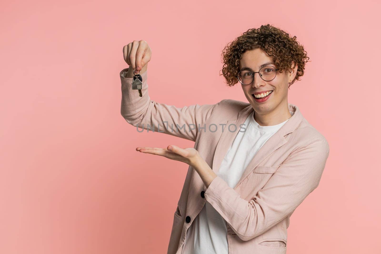 Caucasian young man with curly hair real estate agent lifting hand showing the keys of new home house apartment, buying or renting property, mortgage loan, insurance. Guy isolated on pink background