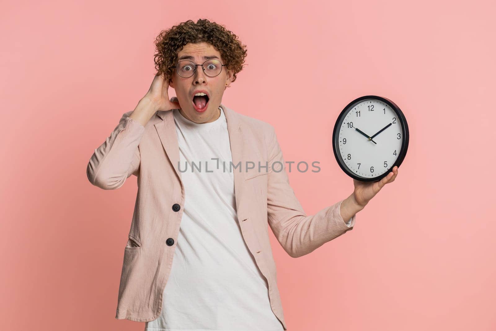 Caucasian curly haired man in jacket with anxiety checking time on clock, running late to work, being in delay, deadline. Young guy looking at hour, minutes, worrying to be punctual on pink background