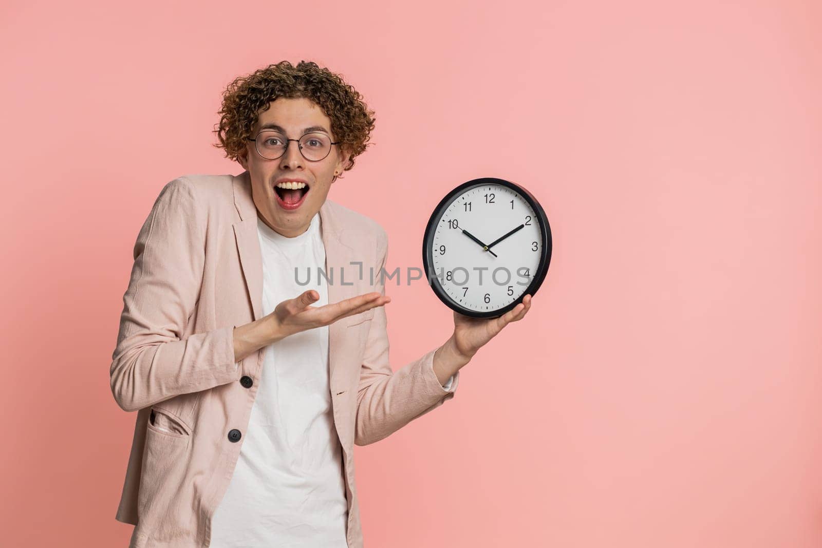 It is your time, hurry up. Caucasian man showing time on wall office clock, ok thumb up, approve, pointing finger at camera, advertisement, deadline. Curly haired young guy isolated on pink background