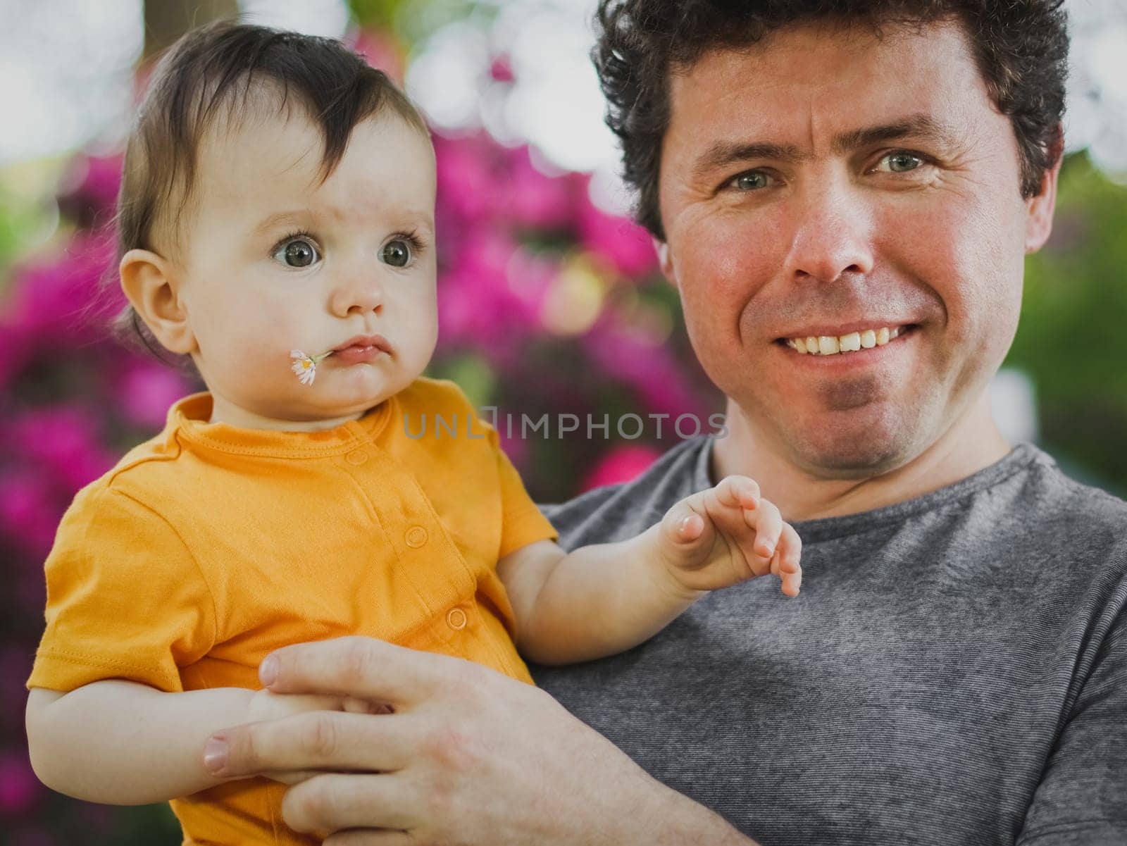 Caucasian young dad man in a gray t-shirt with a happy smile hugs his little daughter in a yellow bodysuit with a daisy in her mouth in a public park, close-up side view. Concept of fatherhood, dads, family vacation, lesson, spring walk, first steps, dad care .