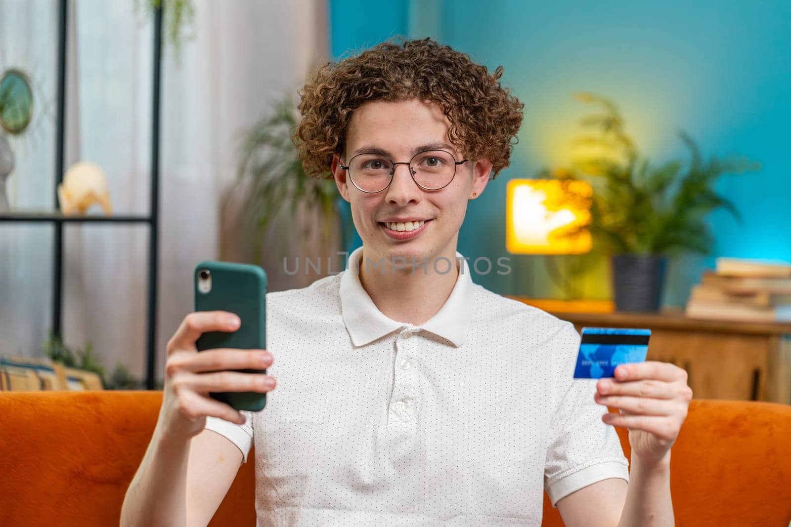 Caucasian man using credit bank card and smartphone while transferring money, purchases online shopping cashless order food delivery at home apartment indoors. Happy young guy sits in room on couch.