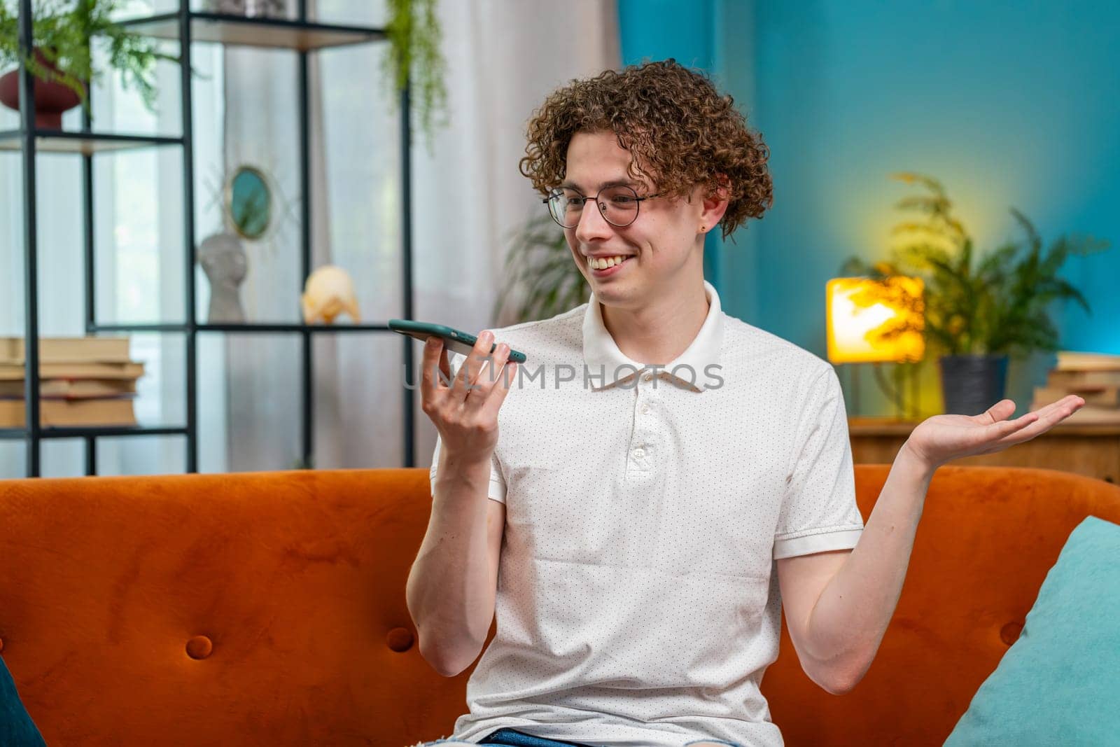 Happy Caucasian man making phone call conversation with friends at modern home room apartment. Young brunette guy enjoying mobile loudspeaker talking gossip, rumors, good news sitting on couch indoors