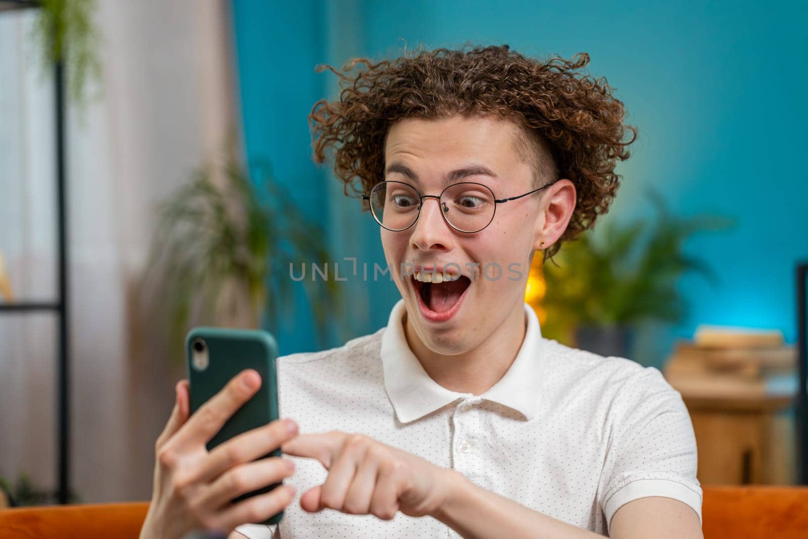 Surprised excited Caucasian young man winner holding smartphone reading good news amazed by online bet bid game win. Happy guy male on sofa looking at screen overjoyed by victory success in apartment.