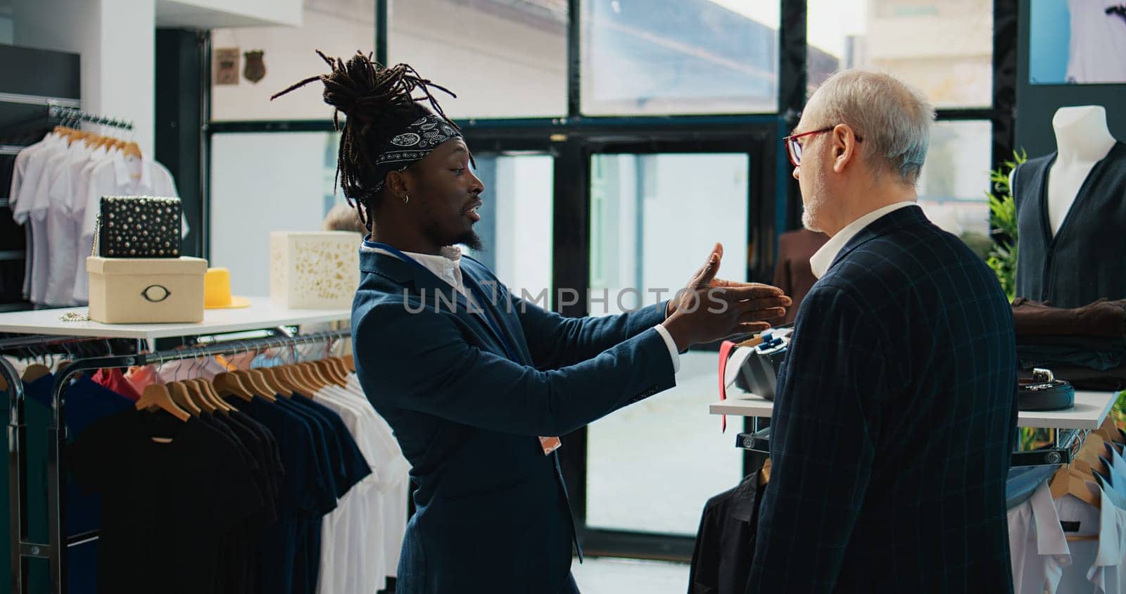 African american worker showing a black shirt and red tie to client, assisting him to buy the perfect outfit for an elegant occasion. Professional stylist gives advice to buyer at store. Camera B.