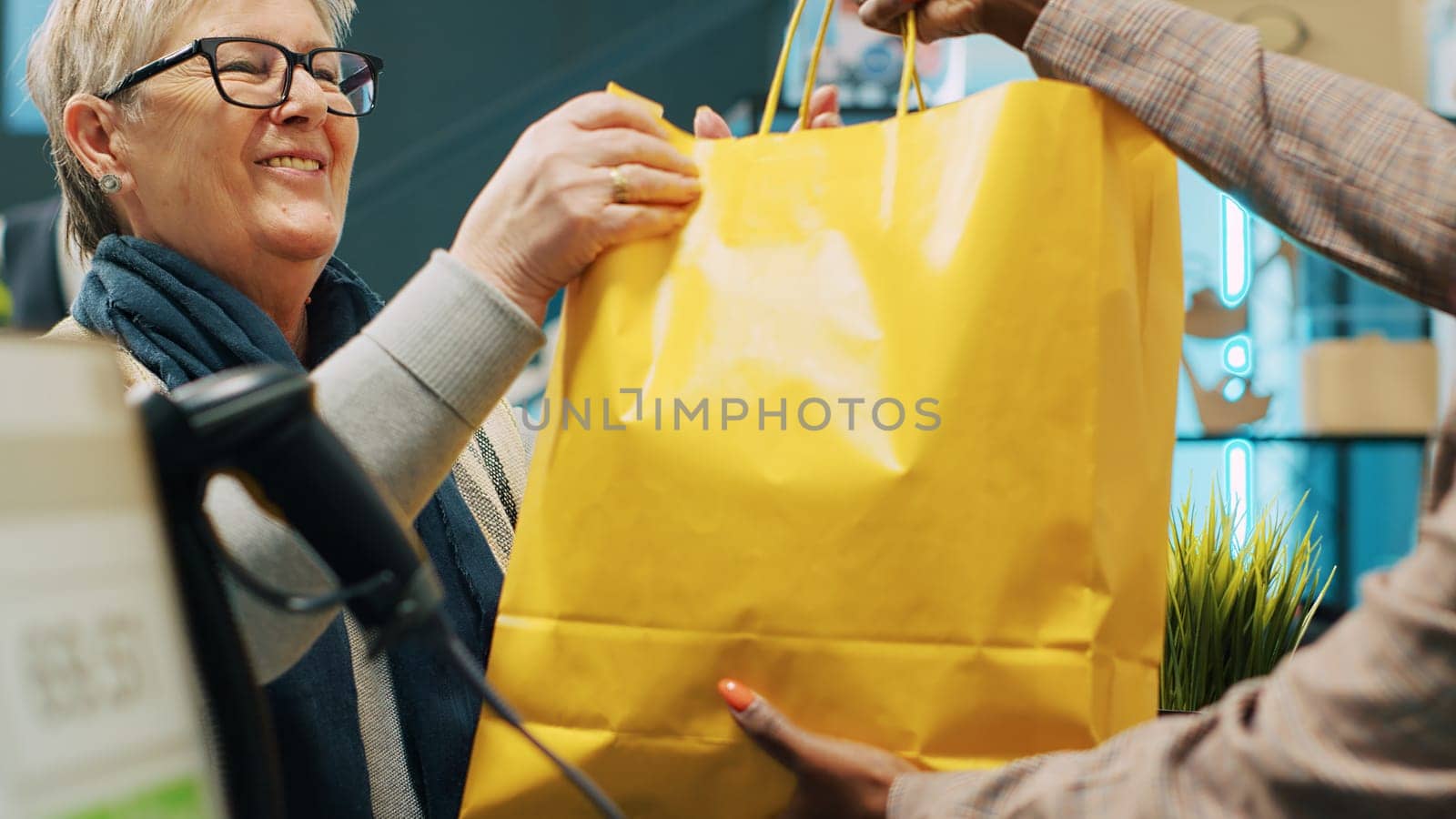 Senior client paying for clothing items at store checkout counter, making card payment at pos terminal to buy clothes from shopping center. African american worker gives bag to shopper. Camera A.