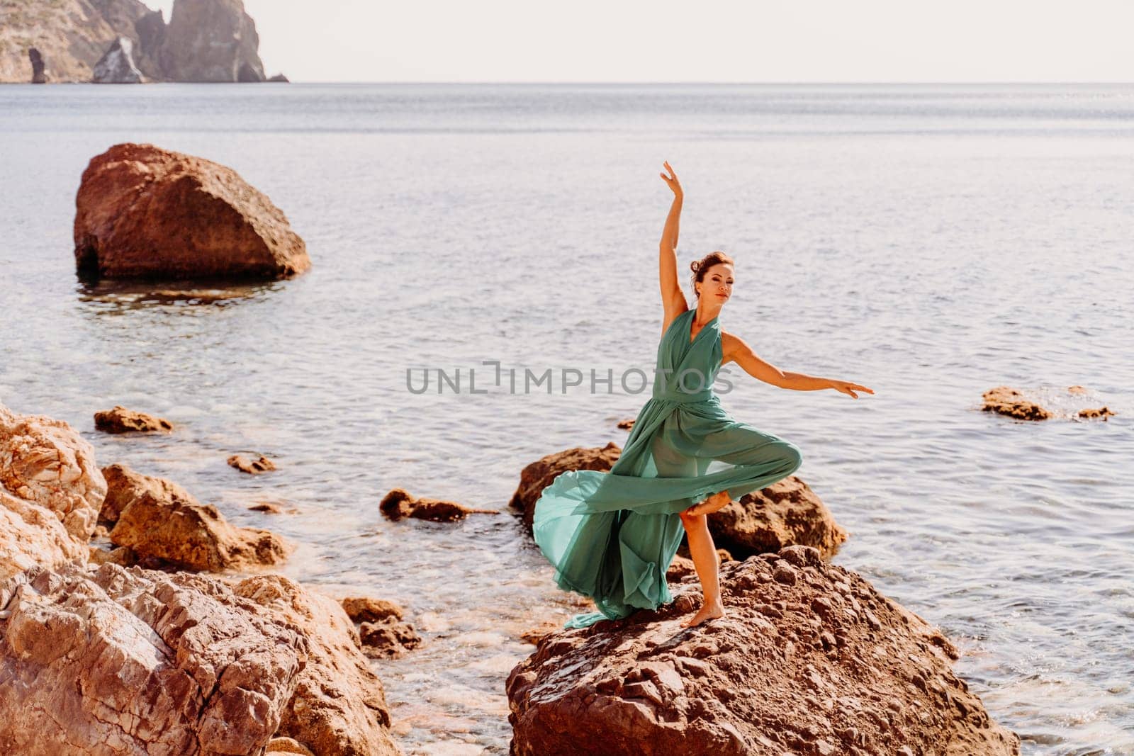 Woman green dress sea. Female dancer in a long mint dress posing on a beach with rocks on sunny day. Girl on the nature on blue sky background