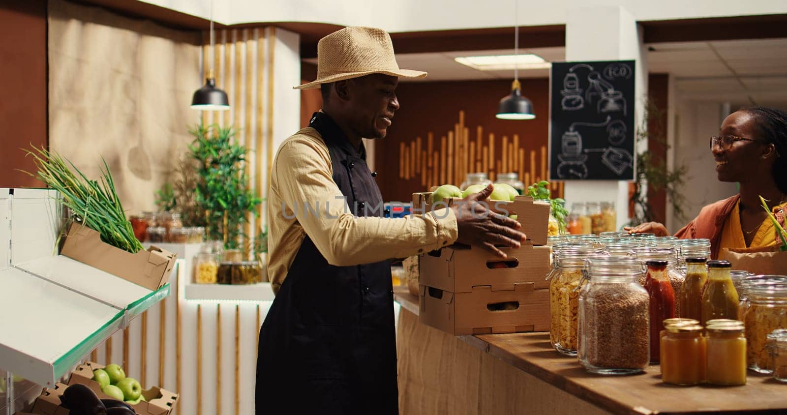 Local vendor placing crates with freshly harvested fruits on grocery store display, recommending organic produce to regular customer. African american farmer restocking all merchandise. Camera 2.