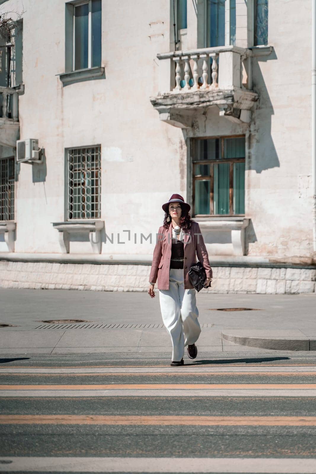 Woman city road crossing. Stylish woman in a hat crosses the road at a pedestrian crossing in the city. Dressed in white trousers and a jacket with a bag in her hands