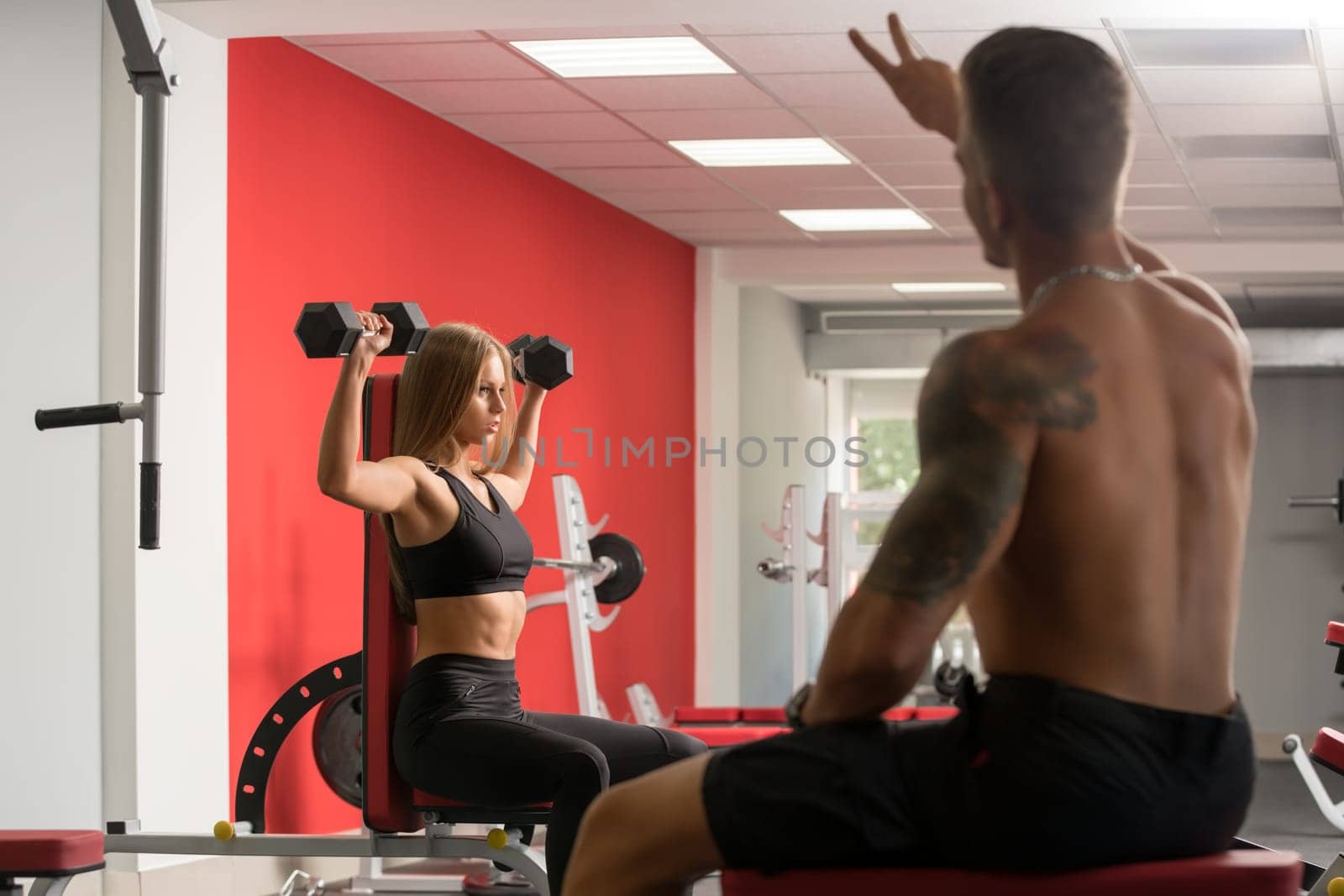 Athletic blonde woman with perfect body in sportswear lifting some weights, working on her biceps with dumbbells in gym, male trainer in foreground