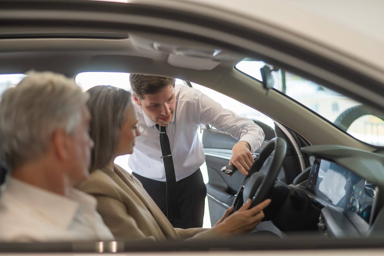 A gray-haired Caucasian woman is sitting behind the wheel of a car. Car dealership salesman consulting mature married couple. by mrwed54