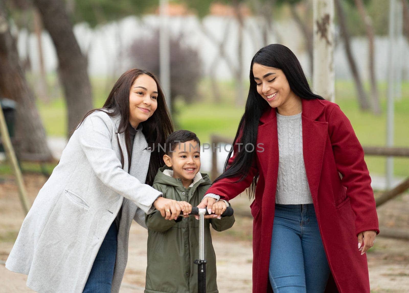 Front view of lesbian latin couple helping their son and teaching how to ride a scooter in a park.LGBT family.