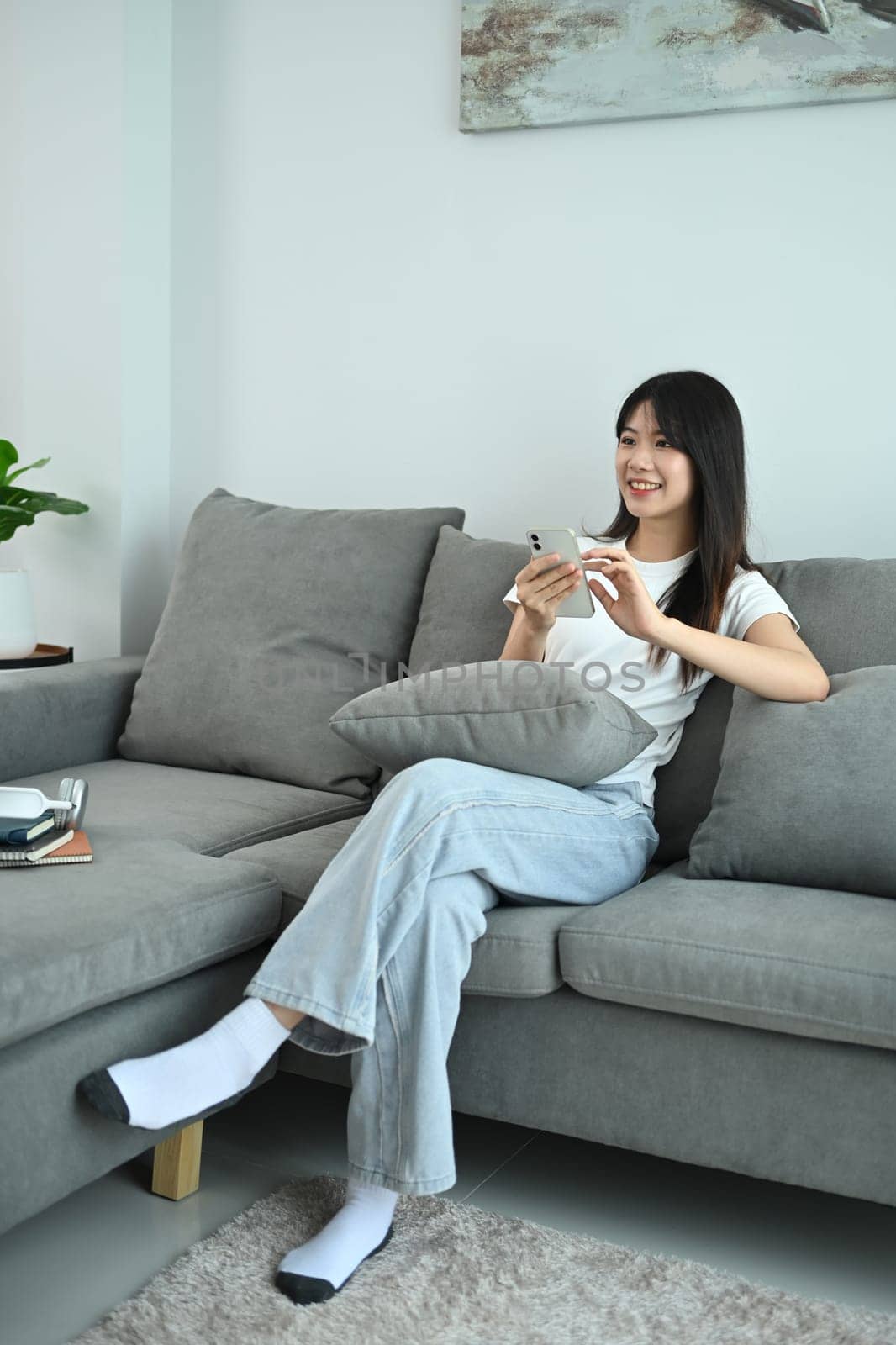 Young woman in casual clothes using mobile phone relaxing on couch at home by prathanchorruangsak