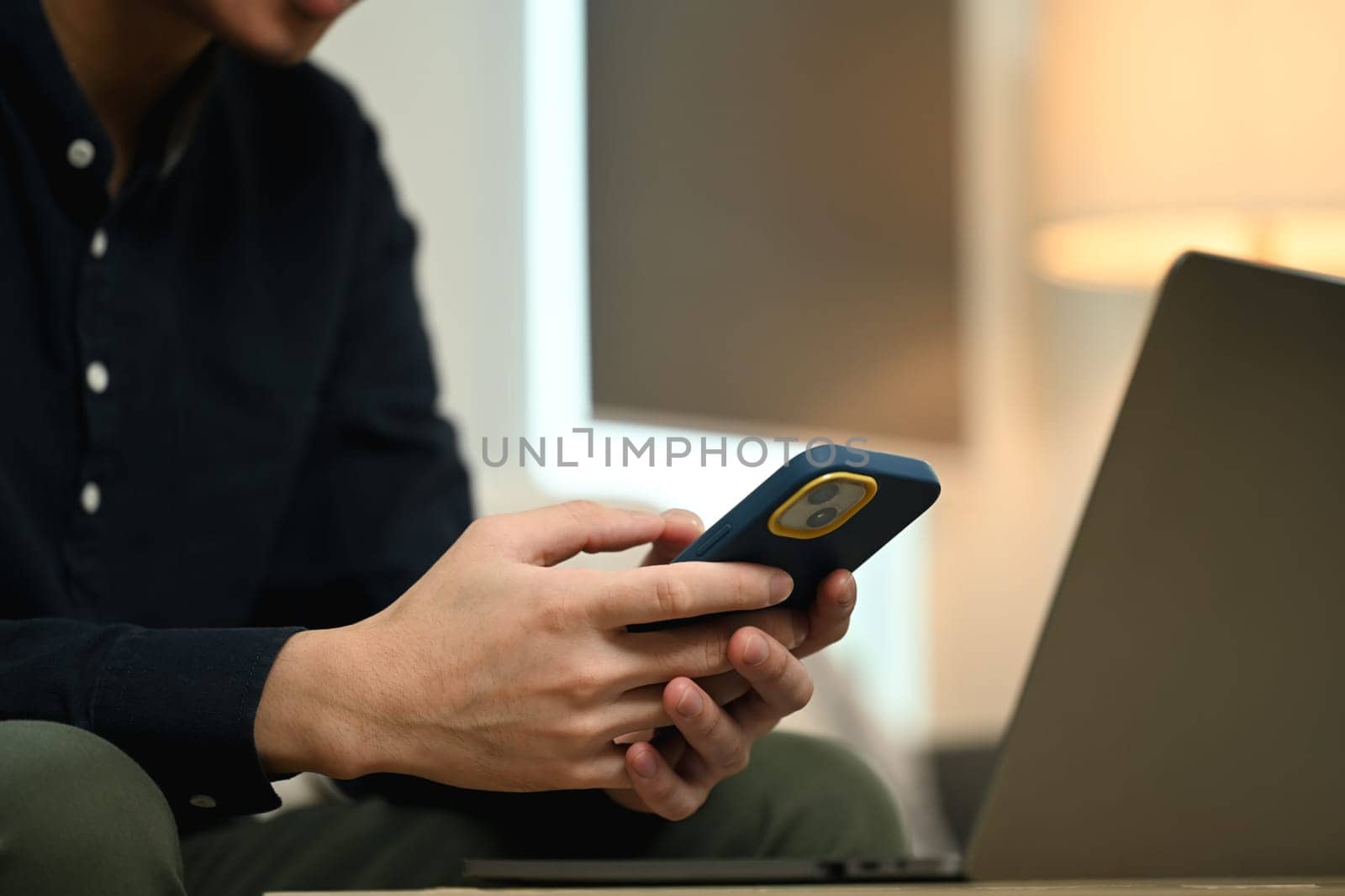 Male freelancer working with laptop and using mobile phone on couch in living room.