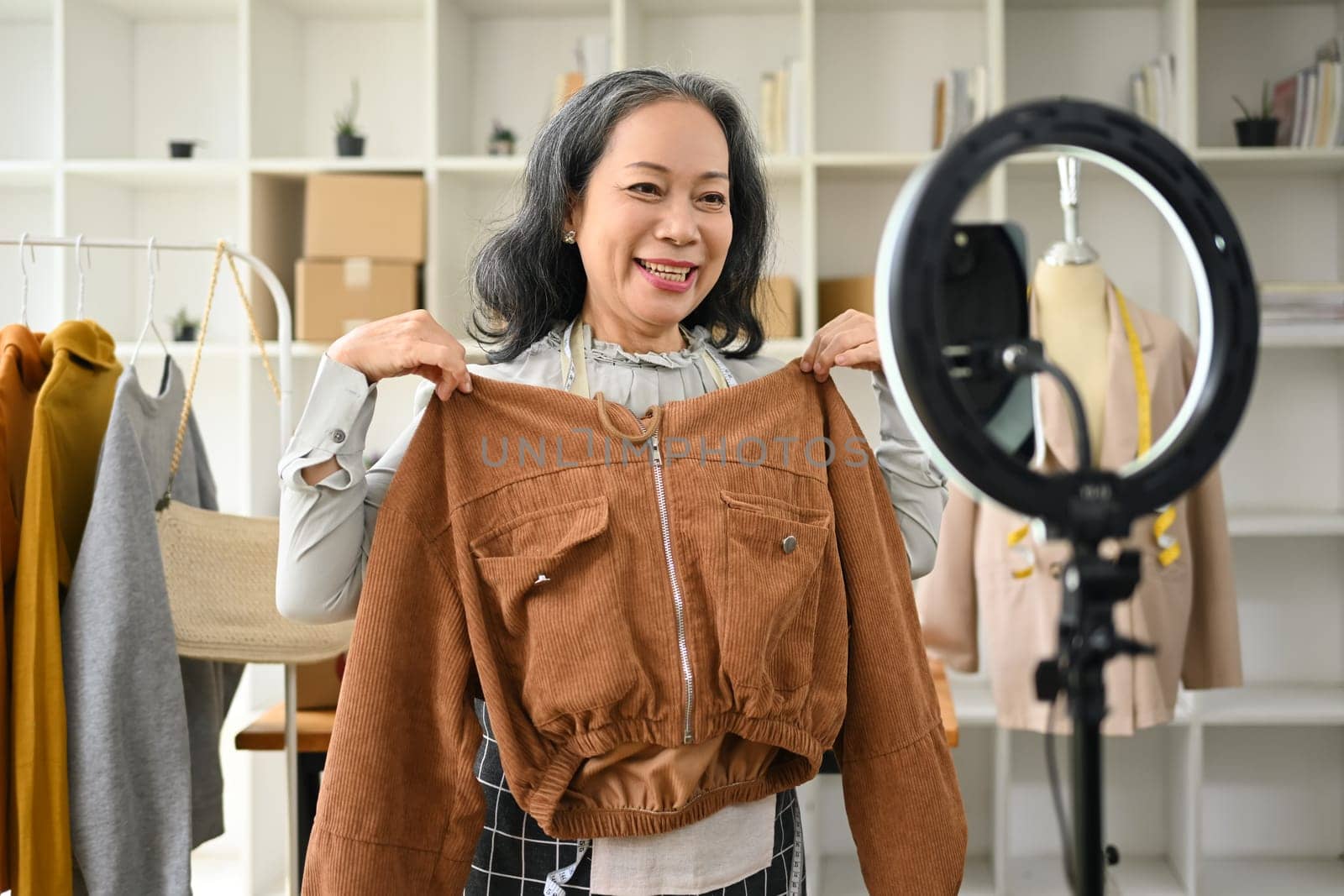 Senior female small business owner showing clothes and present detail during live streaming at her clothing store by prathanchorruangsak