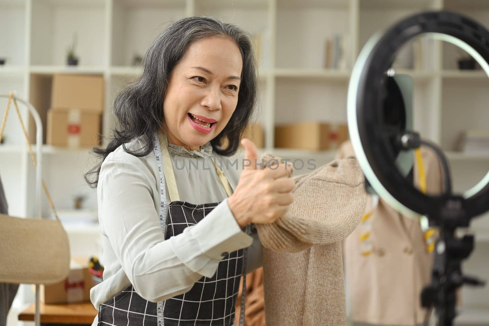 Smiling senior woman presentation bag fashion in front of the camera while live streaming at her shop.