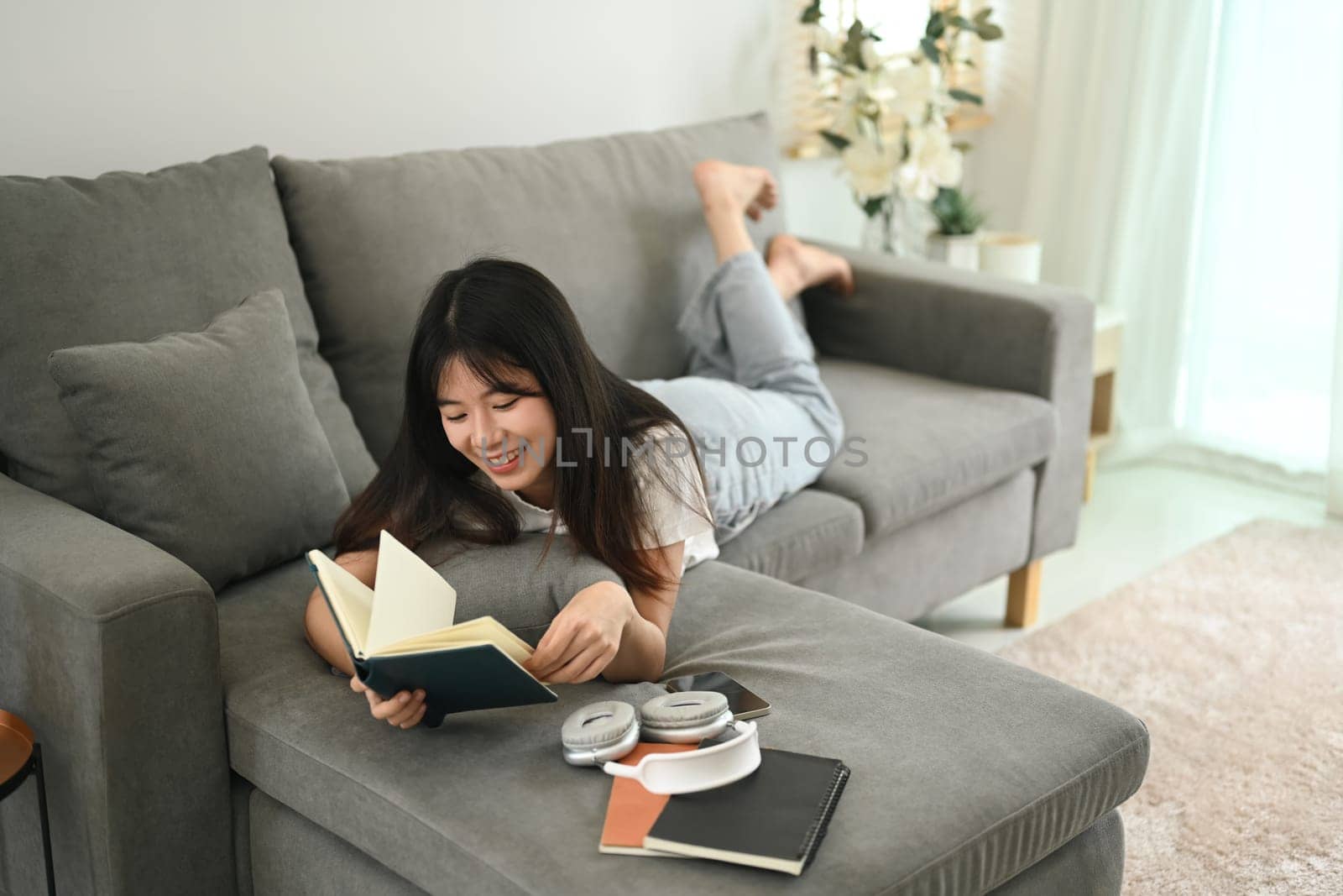 Happy young Asian woman reading book while lying on couch at home.