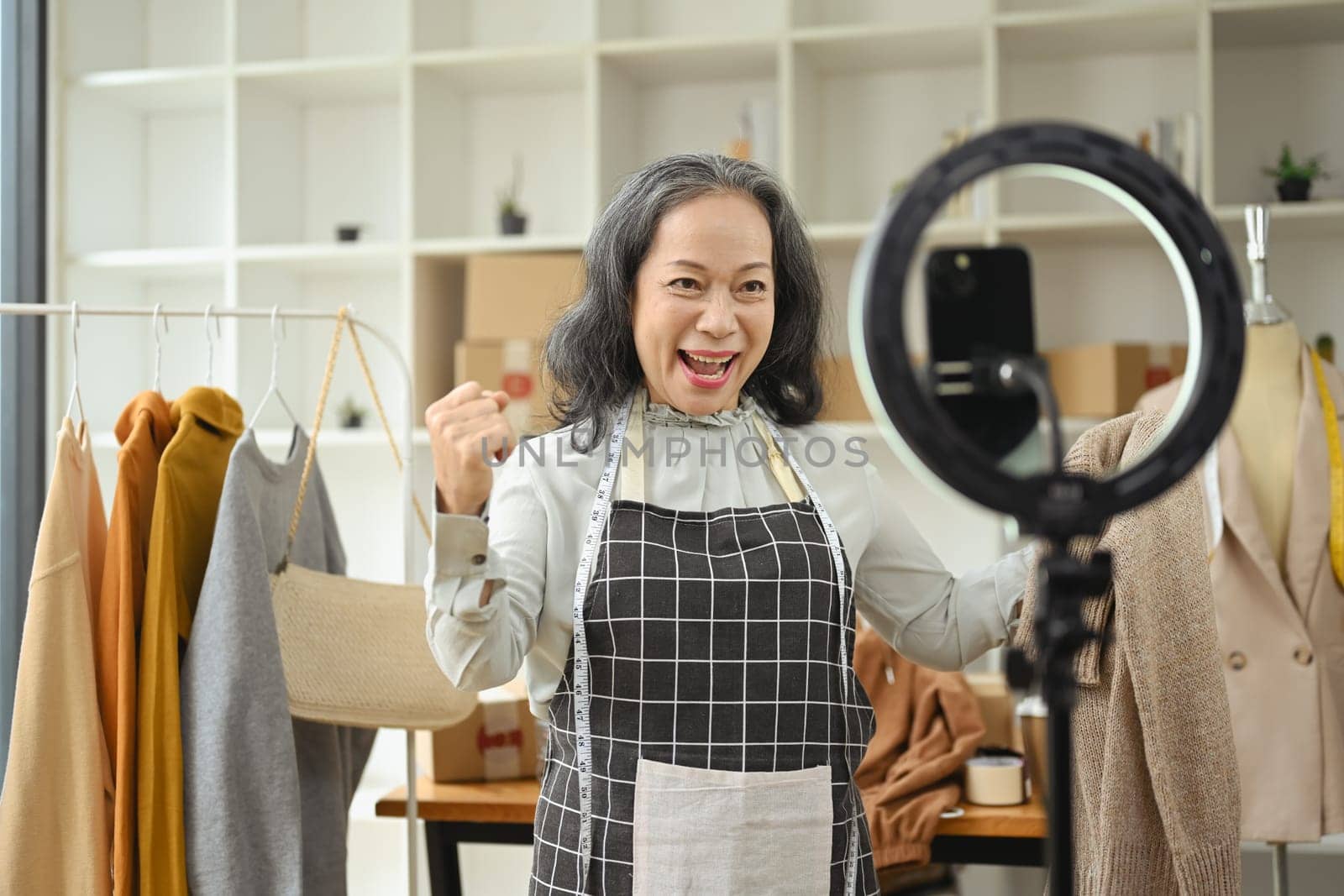 Cheerful senior woman online live stream on mobile phone at her clothing store.